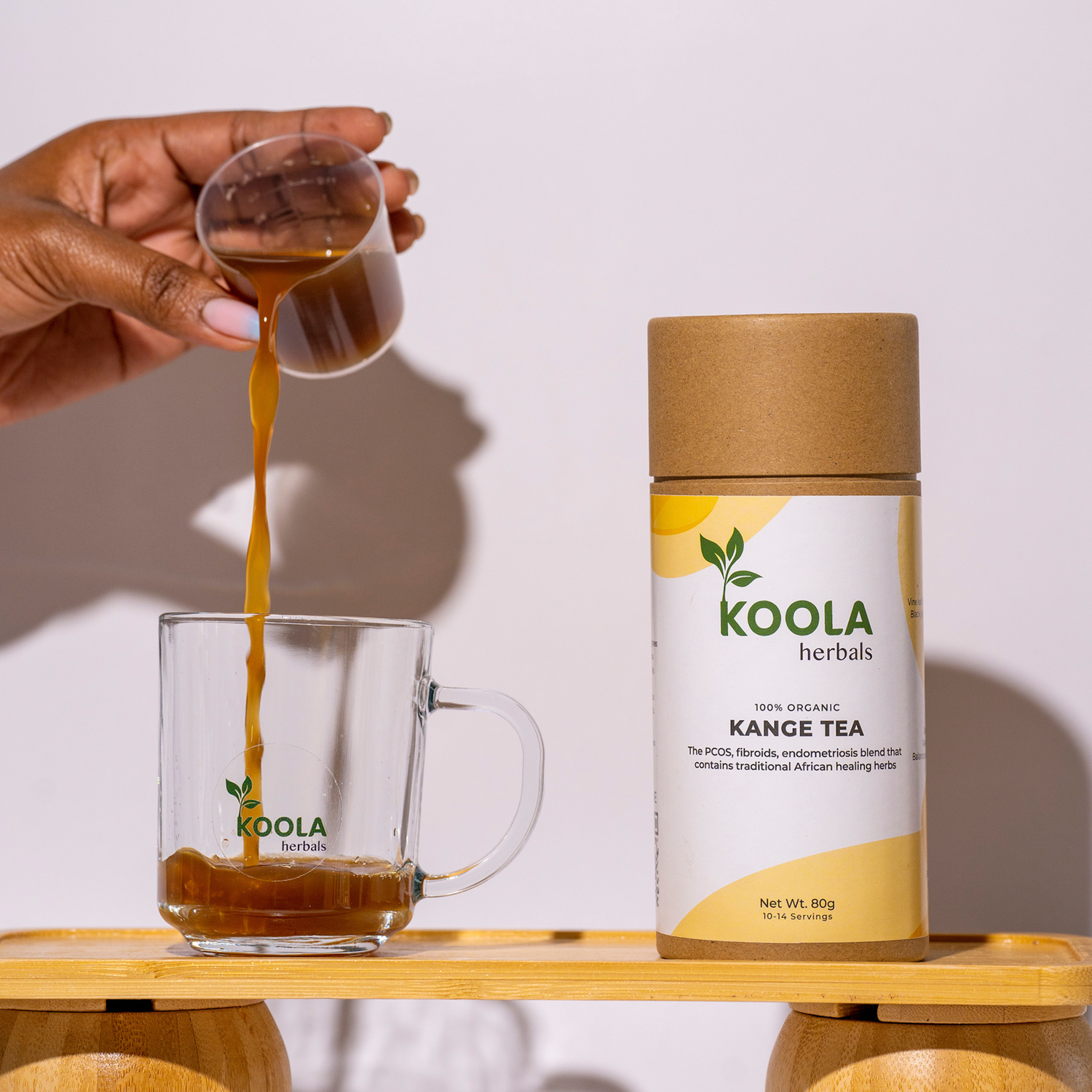 Koola Herbals Kange Tea being poured into a glass with product packaging on a wooden surface.